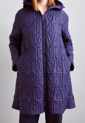 Person iført en lang lilla quiltet frakke med hætte, med broderede bogstaver, der danner ordet "LOVE" i et gentaget mønster.
