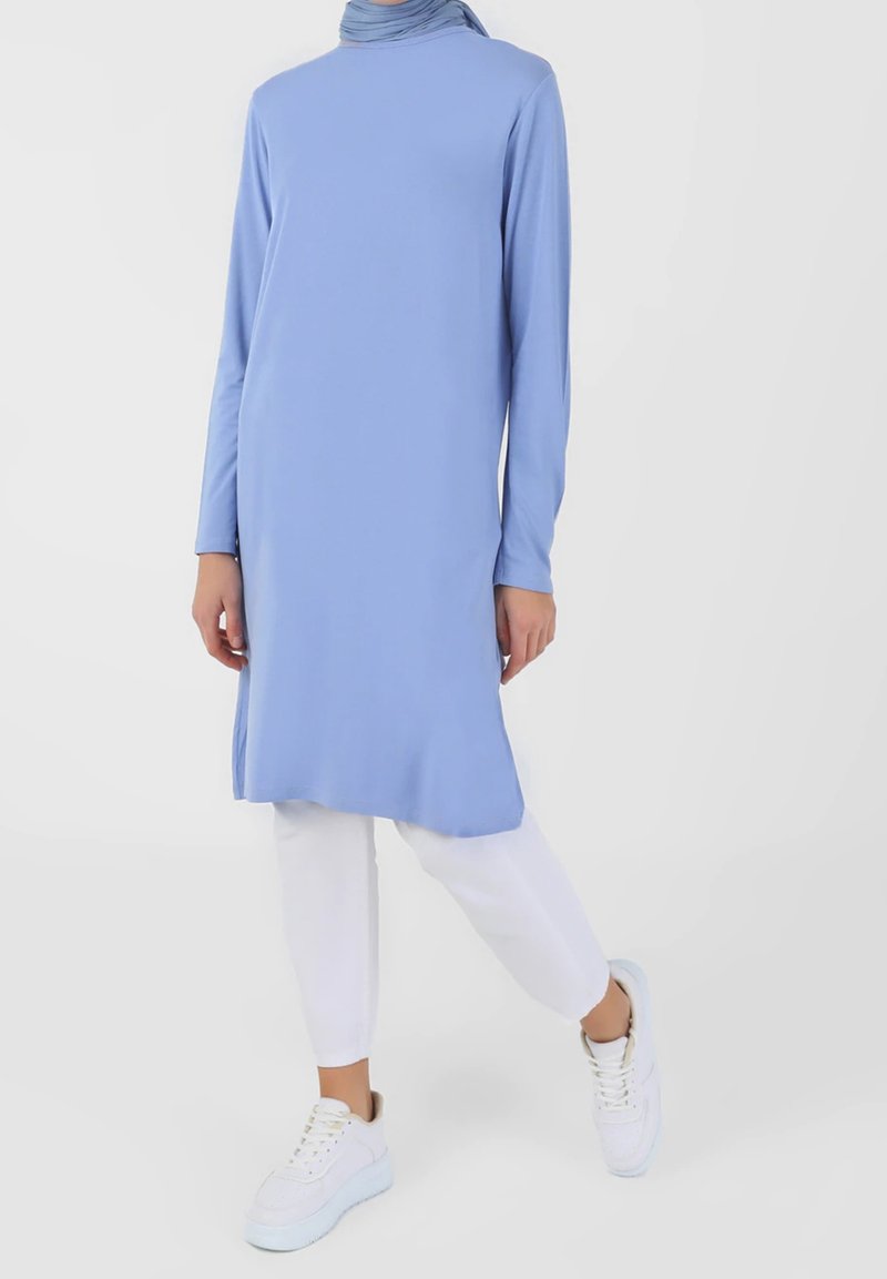 Tunique bleu clair à manches longues avec un col montant, associée à un pantalon blanc coupé au niveau des chevilles et des baskets blanches. Tissu lisse, coupe décontractée, design minimaliste.