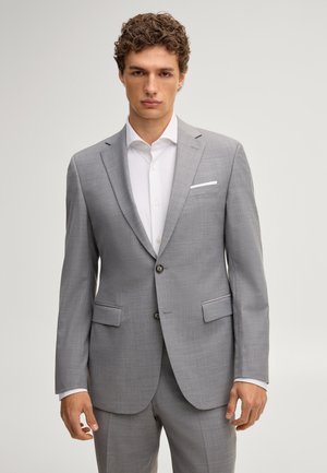 Młody mężczyzna stoi, ubrany w jasnoszary garnitur, białą koszulę i biały poszetkę, neutralne tło.