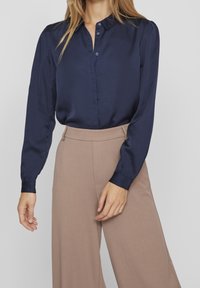 Blusa di raso blu navy con colletto a punta, abbottonatura frontale e maniche arricciate, abbinata a pantaloni a gamba larga beige con vita alta.