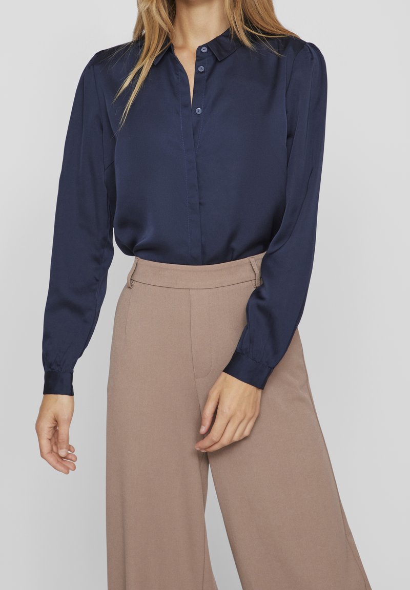 Blusa di raso blu navy con colletto a punta, abbottonatura frontale e maniche arricciate, abbinata a pantaloni a gamba larga beige con vita alta.