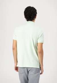 Personne de dos portant un polo vert clair à manches courtes et un jean gris sur un fond blanc uni.