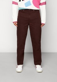 Persona con pantalones rectos de color marrón oscuro y bolsillos delanteros, zapatillas blancas con rayas rojas, y un suéter geométrico colorido.