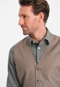 Beige Weste mit glatter Textur, V-Ausschnitt und zwei Knöpfen, getragen über einem grauen Hemd mit strukturiertem Finish.