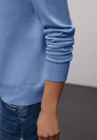 Pull en coton bleu clair avec poignets et ourlet côtelés ; il présente une coupe décontractée avec des manches retroussées, associé à un jean bleu foncé.