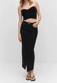 Falda maxi de denim negra con abertura frontal, diseño de cintura alta y bolsillos delanteros. Combinada con un top negro sin tirantes y chanclas negras.