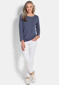 Lächelnde Frau mit langen grauen Haaren trägt ein navyblaues und weiß gestreiftes Langarmhemd, weiße Hosen und weiße Sneakers und steht vor einem einfarbigen Hintergrund.