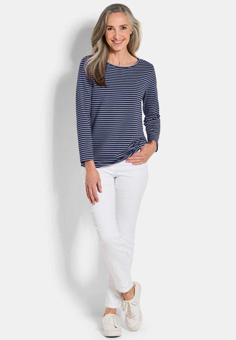 Lächelnde Frau mit langen grauen Haaren trägt ein navyblaues und weiß gestreiftes Langarmhemd, weiße Hosen und weiße Sneakers und steht vor einem einfarbigen Hintergrund.