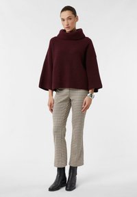 Burgundowa dzianinowa bluzka z dekoltem typu cowl i rękawami dzwonami, w połączeniu z beżowymi i czarnymi spodniami w kratę oraz czarnymi botkami za kostkę.