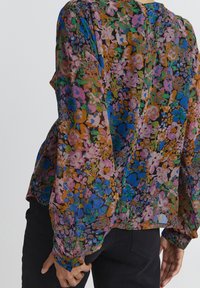 Gennemsigtig blomsterbluse med et livligt mix af pink, blå, grønne og orange blomster; har lange ærmer og en løs pasform. Sorte bukser er synlige.