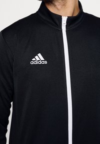 Muž oblečený v černé zipové bundě Adidas s bílým zipem a vyšitým logem na levé hrudi, viditelný od krku po střed trupu.