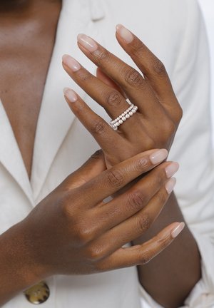 Dark-skinned hands with natural, manicured nails wearing a double-row pearl ring, crossed over the chest in a cream-colored garment.