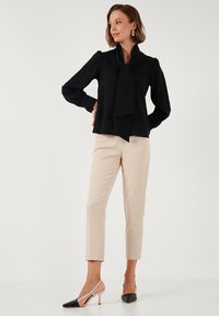 Black long-sleeve blouse with a bow tie detail, paired with beige tapered trousers and black and nude color-blocked heels. Smooth texture.
