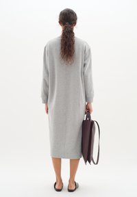 Graues, langärmeliges, knielanges Kleid aus weichem, strukturiertem Stoff. Das Model hält eine braune Ledertasche und trägt schwarze Slipper.