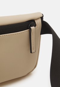 Sac bandoulière beige en matériau lisse avec une fermeture éclair et une sangle noires. Présente des coutures près de la fermeture éclair et un design épuré.