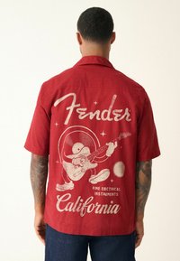 Homme avec des tatouages portant une chemise rouge à manches courtes présentant un graphisme vintage Fender California et un personnage de cartoon jouant de la guitare au dos.