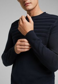 Donkerblauwe gebreide trui met horizontale geribbelde textuur op de schouders, ronde halslijn en aangesloten mouwen met geribbelde manchetten.