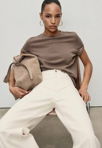 Brown cotton t-shirt paired with white wide-leg jeans. A taupe suede bag with a strap is held in the lap, seated on a chair with a neutral background.