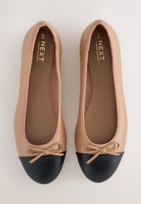 Paire de ballerines beiges avec bouts arrondis noirs et petits nœuds beiges sur un fond clair.