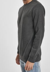 Uomo che indossa un felpa a maniche lunghe grigia scura e jeans azzurri chiari, in piedi contro uno sfondo chiaro e neutro.