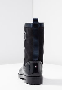 Musta mokkanahkainen naisten saapas, jossa kiiltävä musta kärki, sininen korostusnauha, takana vetokahva ja sivulla logotunnus. Tasapohjainen kumipohja.