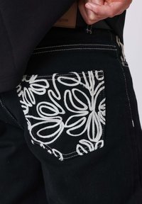 Zwarte broek met een sierlijke zak met witte geborduurde bloemenpatronen, benadrukt door contrasterende stiksels en een zichtbaar taillebandlabel.