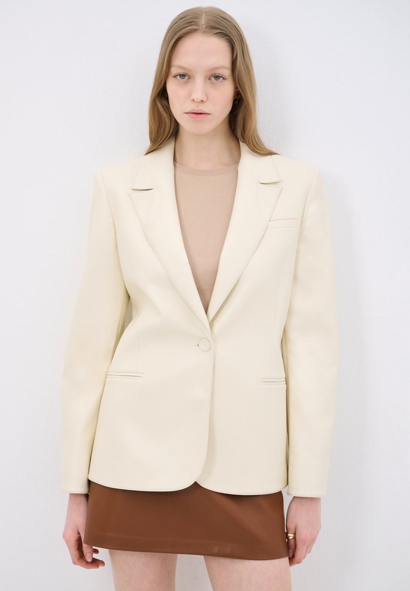 Blazer su misura color crema con un solo bottone, revers a spillo e due tasche frontali, abbinato a una gonna mini in pelle marrone.