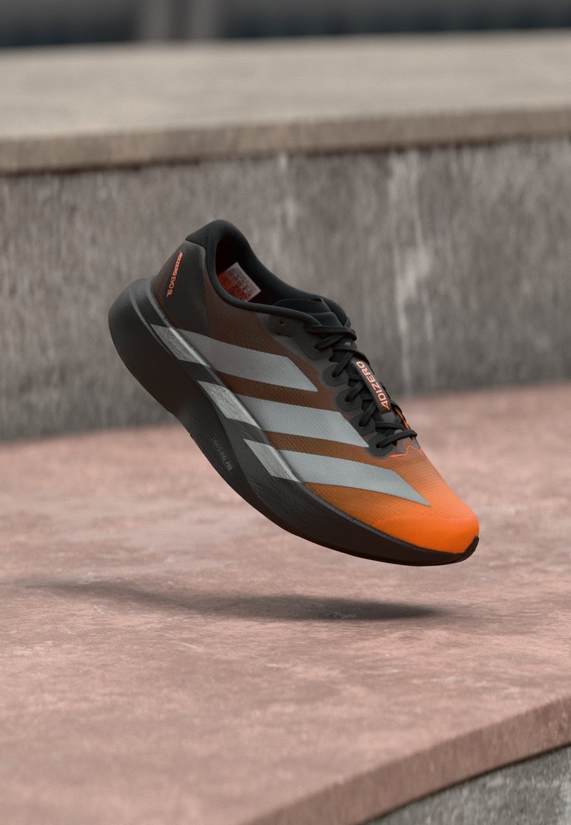 Orangefarbener und schwarzer Sportschuh mit silbernen Streifen und schwarzen Schnürsenkeln, mit der Aufschrift "ADIZERO", der über einer Betonfläche zu schweben scheint.