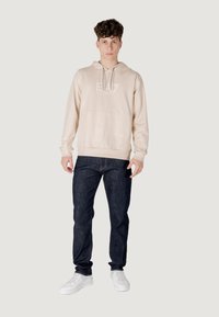 Felpa beige con tasca frontale e cappuccio con lacci, abbinata a jeans in denim scuro e sneakers bianche. La felpa presenta una grafica con logo.