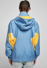 Giacca azzurra chiara con dettagli gialli e bianchi, dotata di cappuccio, polsini elasticizzati e vestibilità comoda. Realizzata in un materiale morbido e lucido.