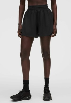 Person iført sorte atletikshorts med elastik i taljen, sorte sokker og sorte løbesko, står mod en ensfarvet baggrund.