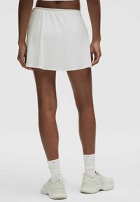 Falda deportiva blanca con una textura suave, de longitud a medio muslo y cintura elástica. Combinada con zapatillas deportivas blancas y calcetines.