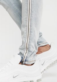 Hellblaue Jeanshose mit seitlichem Reißverschluss und silbernem Zubehör, kombiniert mit weißen Nike Air Max Sneakern.