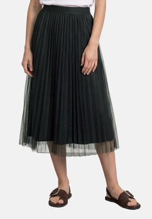Femme portant une jupe midi plissée noire avec un voile transparent, un haut blanc et des sandales ouvertes marron foncé avec de grandes boucles.