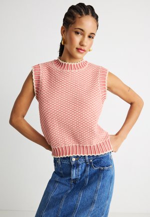 Woman with braided hair wears sleeveless pink knit top and high-waisted blue denim skirt, posing with hands on hips against plain background.