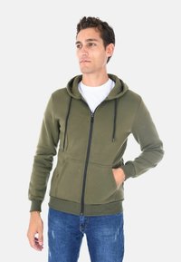 Felpa con zip di colore verde oliva, con chiusura frontale a zip, cappuccio con cordini e due tasche laterali. Tessuto morbido con polsini e orlo a coste.