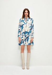 Blumiges Mini-Kleid mit weißem Hintergrund, blauen Blättern und cremefarbenen Blumen. Verfügt über einen Kragen, eine Knopfleiste und einen gerafften Saum. Getragen mit beigen kniehohen Stiefeln.