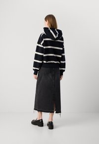 Hoodie a righe bianco e nero con tasca frontale e cappuccio regolabile, abbinato a una lunga gonna di jeans nera con spacco posteriore e orlo sfilacciato.