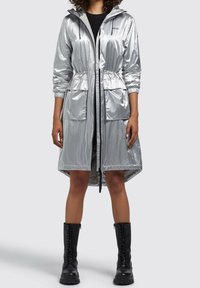 Srebrny płaszcz przeciwdeszczowy z kapturem, z wąskim pasem, dwoma przednimi kieszeniami i elastycznymi mankietami. Nosi się go na czarnej sukience, w zestawieniu z czarnymi bojówkami.