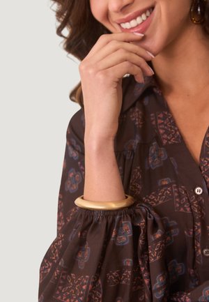 Camisa marrón con estampado, con mangas abullonadas elásticas y botones, complementada con una pulsera de bangle lisa de color dorado en la muñeca.