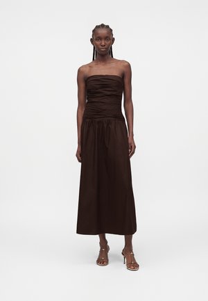 Punase palmikutega naine, kes kannab tumepruuni rihmadeta voltidega midi-kleit ja rihmikuid kõrgete kontsadega sandaale ühtlasel taustal.