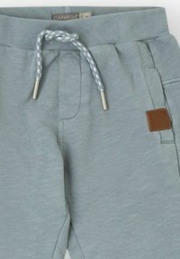 Pantalones de chándal gris claro con cintura acanalada, cordón ajustable y una etiqueta de cuero marrón en el lateral. Cuenta con bolsillos laterales.