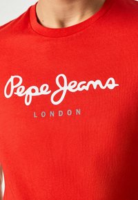 Camiseta de algodón rojo con el logotipo en cursiva blanca "Pepe Jeans" y el texto más pequeño en gris "LONDON". Textura suave con diseño de cuello redondo.