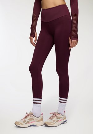 Leggings bordeaux con vita alta, realizzati in materiale morbido, abbinati a scarpe da ginnastica beige e rosa e calze bianche con strisce marroni.