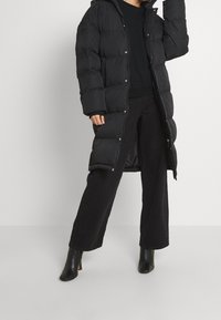Abrigo de plumas largo y negro con capucha, textura acolchada, cierre de botones a presión, llevado sobre un suéter negro y pantalones de pierna ancha, combinado con botas de tacón negras.
