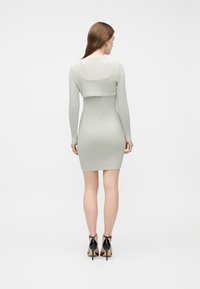 Γυναίκα με μακριά καστανά μαλλιά που φοράει εφαρμοστό μακρυμάνικο φόρεμα σε ανοιχτό γκρι και μαύρες ψηλοτάκουνες γόβες, γυρισμένη πλάτη σε απλό φόντο.