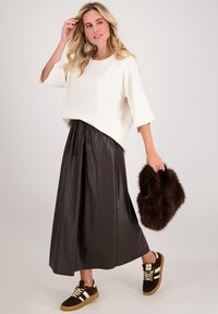 Crème-kleurige trui met driekwart mouwen, gecombineerd met een vloerlange bruine faux leren rok en bruine sneakers met witte strepen. Houdt een bruine bont clutch vast.
