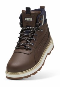 Bota alta de Puma marrón con cordones de patrón negro y blanco, cuello acolchado en azul marino y suela gruesa blanca con pisada marrón.