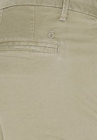 Ljust beiga byxor med en slät textur, med en bakficka och subtil logobroderi. Skräddarsydd design och platta sömmar är synliga.