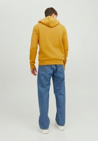 Felpa gialla con cappuccio posteriore, polsini a coste, abbinata a jeans in denim blu e sneakers bianche, vista da dietro.